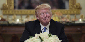 Ein Mann mit orange-blonden Haaren sitzt hinter einem weißen Blumenstrauß und lächelt