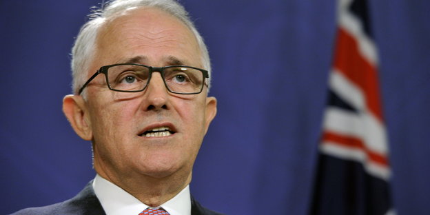 Ein Mann mit gruane Haaren und Brille spricht, im Hintergrund eine australische Flagge
