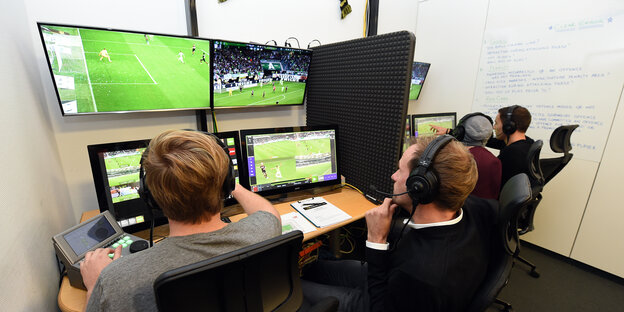 Zwei Männer sitzen mit Kopfhörern vor mehreren Fernsehbildschirmen, auf denen Fußball zu sehen ist