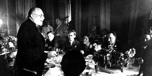 Ein schwarz-weiß Foto zeigt mehrere Männer, die um einen Tisch sitzen, einer von ihnen steht