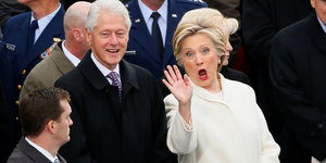 Hillary und Bill Clinton stehen nebeneinander. Hillary winkt und verzieht das Gesicht