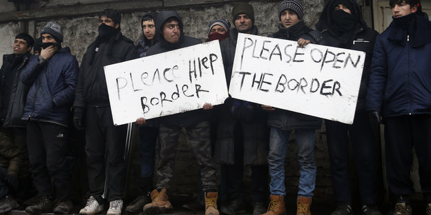 Männer stehen draußen und halten Schilder hoch. Auf einem steht: „Please open the border".