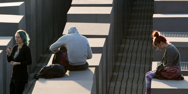 drei Menschen sitzen, jeder für sich, auf Stelen des Denkmals für die ermordeten Juden Europas