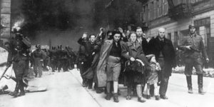 Mit erhobenen Händen und zusammengedrängt geht eine Gruppe polnischer Juden - von SS-Schergen flankiert