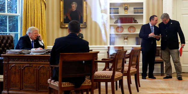 Donald Trump an seinem Schreibtisch im Oval Office. Er telefoniert, während Mitarbeiter sich beraten