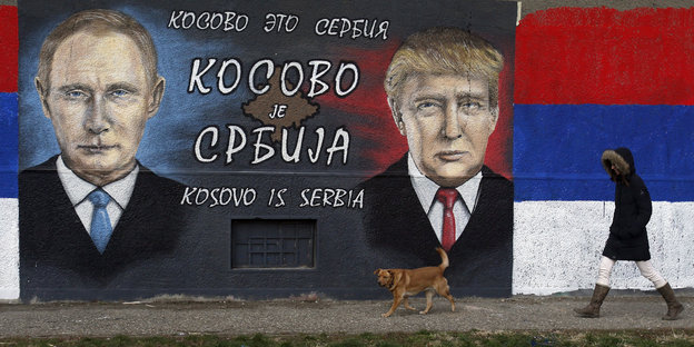 Eine Frau und ein Hund laufen an einem Graffiti von Trump und Putin vorbei