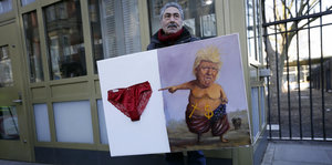 Ein mann hält ein Kunstwerk auf dem ein nackter Donald Trump auf eine Unterhose zeigt