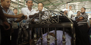 Der Leichnam von Ko Ni liegt bei der Beerdigung in einem Sarg, der aussieht wie ein Bett, das mit verzierten Metallstäben geschmückt ist. Um ihn herum fotografieren viele Männer den Toten.