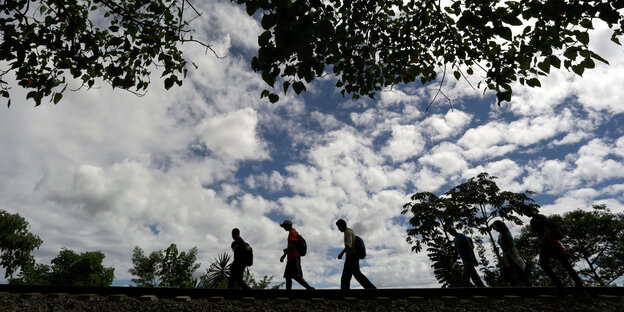 Sechs Menschen laufen hintereinander Gleise entlang, über ihnen erstreckt sich ein Himmel mit Wolken