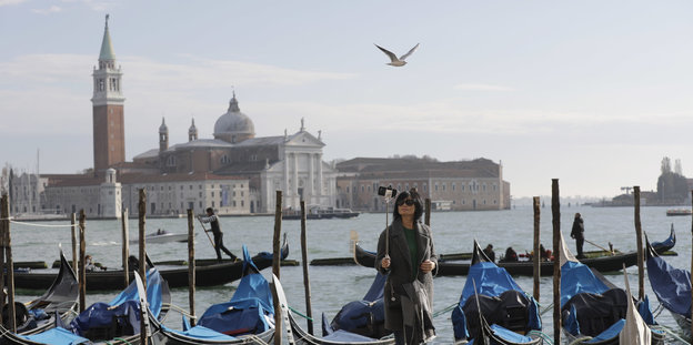 Eine Frau mit seinem Selfiestick steht in Venedig am Wasser, hinter ihr Boote