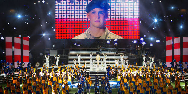 Auf einer Riesenleinwand ist ein Soldat zu sehen, darunter tanzen Cheerleader