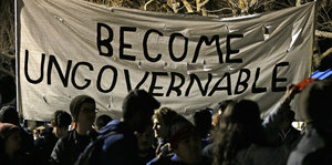 Menschen stehen bei schwacher Beleuchtung beieinander. Auf einem Banner steht: Become Ungovernable