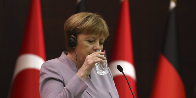 Angela Merkel hebt ein Glas Wasser zum Mund, hinter ihr die Fahnen der Türkei und Deutschlands