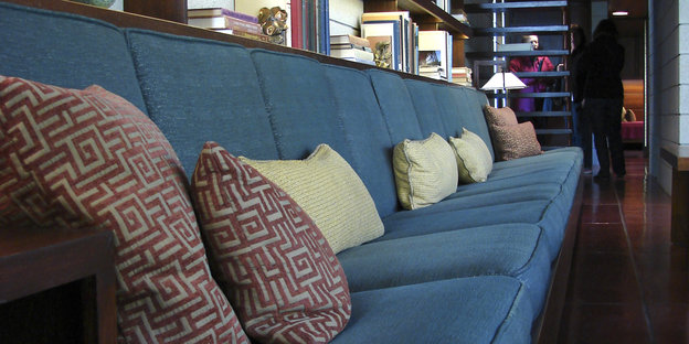 Man sieht eine lange blaue Couch mit vereinzelten beigen und dunkelroten Kissen darauf.