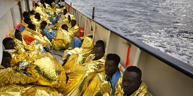 Mehrere Menschen mit Goldfolie umhüllt kauern an Deck eines Schiffes