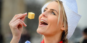 Eine Frau mit weißer Haube hält sich ein Stück Käe aufgespießt auf einen Zahnstocker vor den Mund