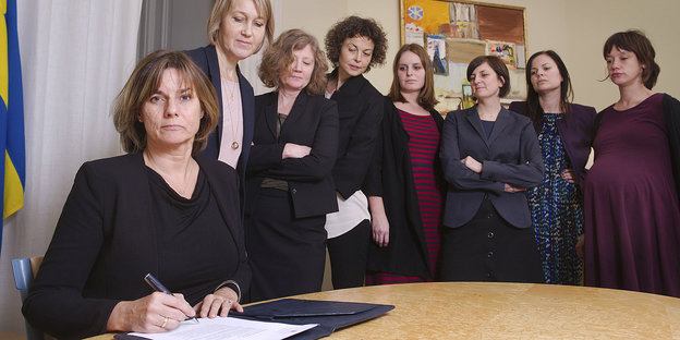 Eine sitzende Frau unterzeichnet ein Dokument, andere Frauen stehen hinter ihr. Alle blicken ernst in die Kamera