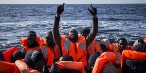 Ein Boot voller Menschen auf dem Meer, in der Mitte hält ein Mann beide Arme gen Himmel
