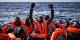 Ein Boot voller Menschen auf dem Meer, in der Mitte hält ein Mann beide Arme gen Himmel