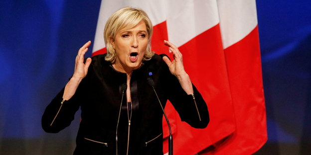 eine Frau mit erhobenen Händen und offenem Mund vor einer französischen Fahne