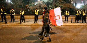 Ein Mann geht mit einem Kind eine Straße an Polizisten vorbei entlang und trägt eine polnische Flagge