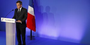 Ein Mann in Anzug steht an einem Rednerpult mit Mikrofon, hinter ihm steht die französische Nationalflagge