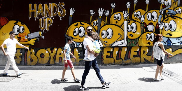 Graffiti in Istanbul: Gelbe Smileys werden bedroht und halten die Hände hoch. Vor der Wand läuft ein Mann mit einem Kind im Arm, der in die Kamera grinst.