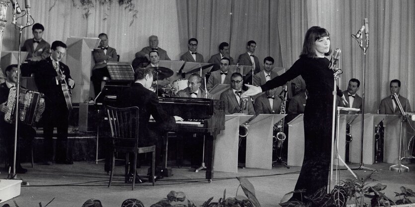 Ein schwarz-weiß Bild, auf dem eine Frau in einem schwarzen Kleid an einem Mikrofon sing und eine Instrumental-Band im Hintergrund spielt