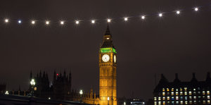 Westminster-Palast bei Nacht, Big Ben ist erleuchtet, eine Lichterkette zieht am Bildrand entlang