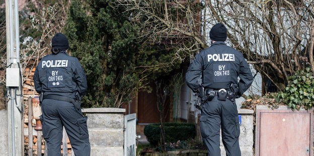 Zwei Polizeibeamte stehen mit dem Rücken zum Betrachter vor einem von Sträuchern zugewucherten Haus
