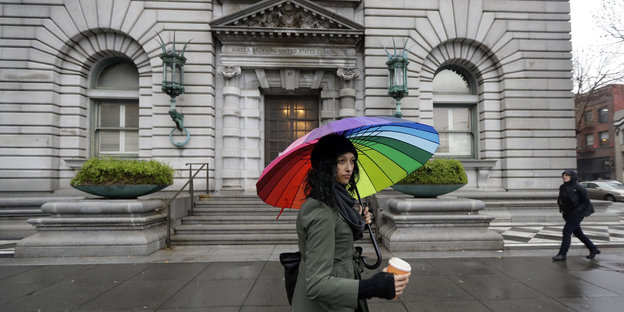 Eine Frau mit einem bunten Regenschirm läuft vor einem grauen Gebäude entlang