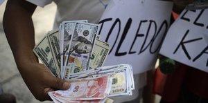 Ein Mensch hält Imitate von Geldscheinen in seiner Hand, dahinter ist ein Schild mit der Aufschrift „Toledo“ zu sehen