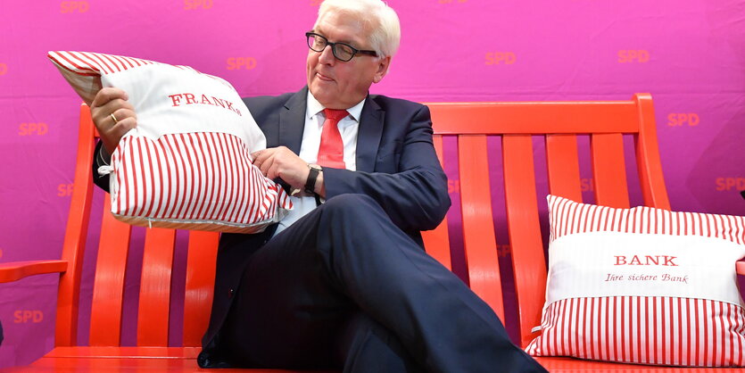 Frank-Walter Steinmeier sitzt mit zwei Kissen, auf denen "Franks Bank" steht, auf einer roten Bank