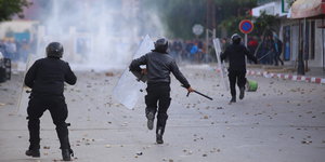 Drei Polizisten rennen auf einer weitgehend leeren Straße Demonstranten hinterher, im Hintergrund steigt Rauch auf