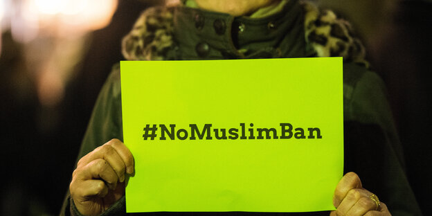 Frau hält gelbes Schild hoch mit Hashtag #NoMuslimBan
