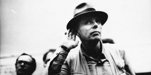 Joseph Beuys in Weste und mit typischem Hut hält sich süffisant die Hand ans rechte Ohr