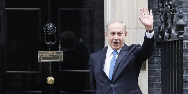 Benjamin Netanjahu steht vor einem Haus und winkt mit der linken Hand