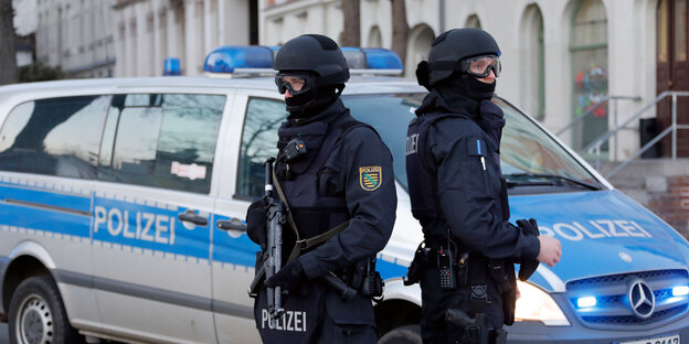 Zwei Polizisten in voller Schutzmontur stehen Rücken an Rücken vor einem Polizeiauto