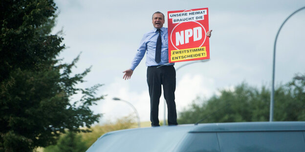 Ein Mann steht auf einem Autodach und hält ein NPD-Plakat im Arm