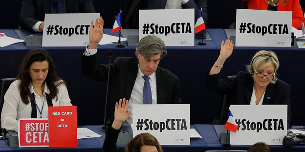 Mehrere Menschen sitzen mit "Sotp Ceta"-Schildern an Tischen und heben die Hände