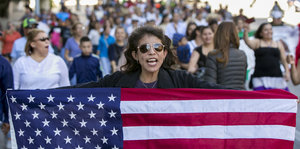 Eine Frau, die eine Sonnenbrille trägt, hält eine US-Fahnen in ihren Händen. Hinter ihr laufen Menschen
