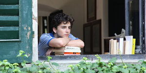 Ein junger Mann blickt, auf einen Bücherstapel gestützt, verträumt aus einem Fenster