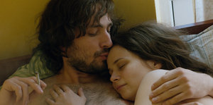 Ein Mann und eine Frau liegen nackt in einem Bett, er küsst sie auf die Stirn und hält eine Zigarette in der Hand