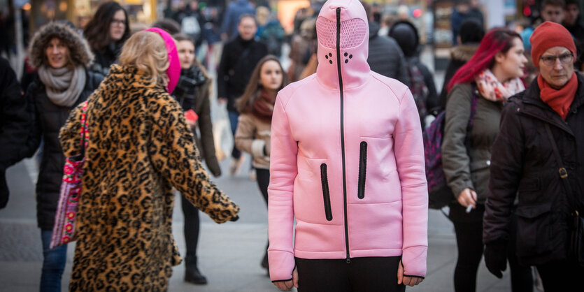 Eine Person trägt in einer Fußgängerzone einen rosafarbenen gummiartigen Kapuzenpullover, der das Gesicht maskiert.