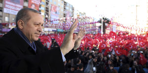 Erdogan grüßt mit versteinerter Miene, im Hintergrund schwenken Anhänger die türkische Flagge
