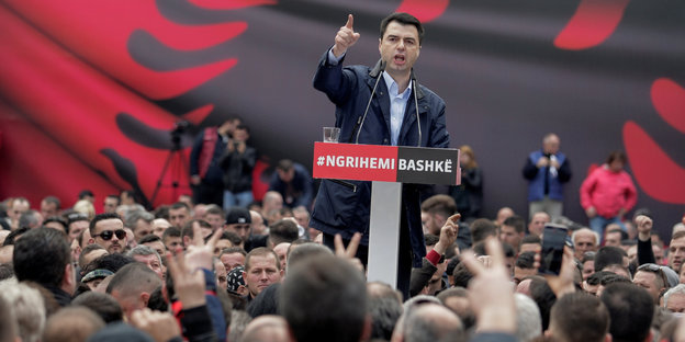 Im Hintergrund eine übergroße rote Flagge mit einem schwarzen Adler, im Vordergrund ein Mann vor einem Rednerpult inmitten einer Menschenmenge