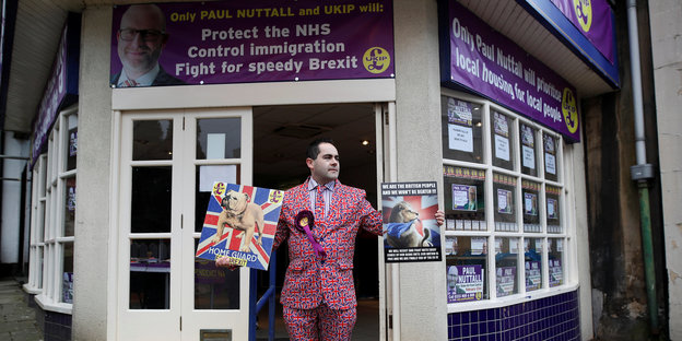 Ein Mann in einem Anzug, auf dem lauter britische Nationalflaggen abgedruckt sind steht vor einem Haus an einer Straßenecke