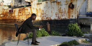 Ein Migrant sitzt auf einem Steg und hält ein Handy in den Händen, hinter ihm liegt ein rostiges Schiff im Wasser