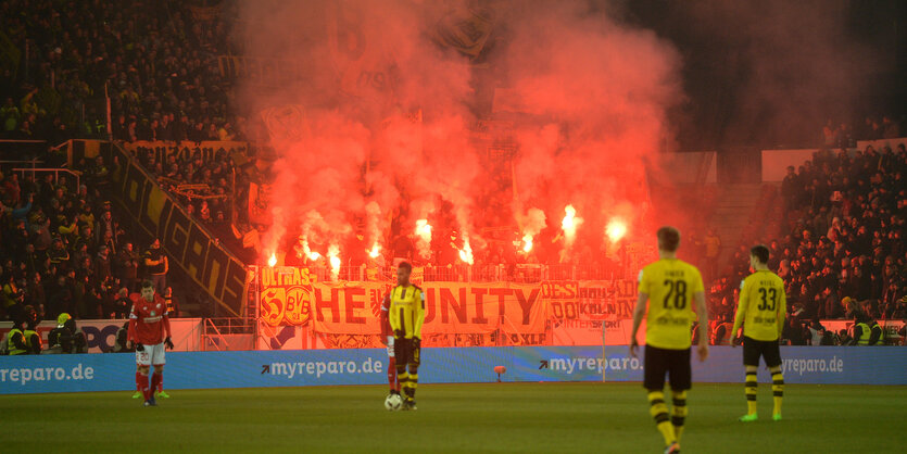 Fußballspieler in gelben Trikots stehen auf dem Fußballfeld, auf der Tribüne sind mehrere Feuer gezündet