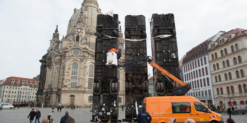 Drei senkrecht aufgestellte Busse vor einer Kirche, ein orangenes Auto mit Personenkran davor